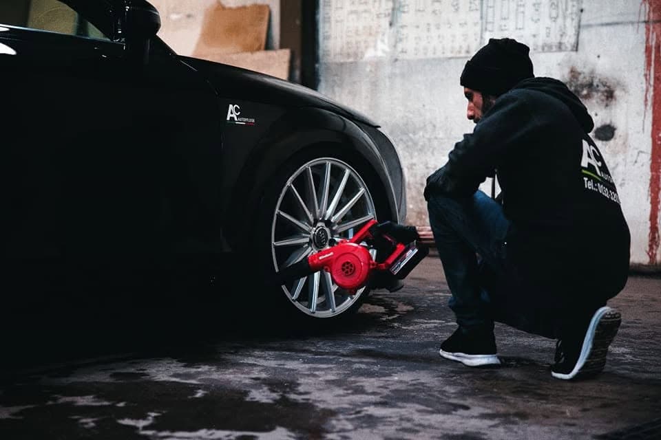 Der Inhaber im AC-Autopflege-Hoodie trocknet in der abgedunkelten Werkstatt die Felge eines schwarzen Fahrzeugs mit einem Akku-Gebläse.